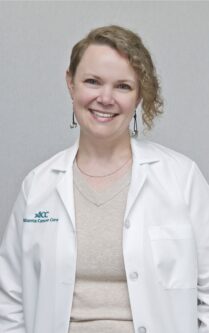 Smiling female clinician in a white lab coat with the Atlanta Cancer Care logo, posed against a gray backdrop. 