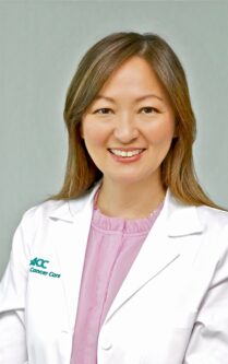 Smiling woman in a white lab coat with NCC Cancer Care logo, pink blouse, portrait photo, professional medical provider. 