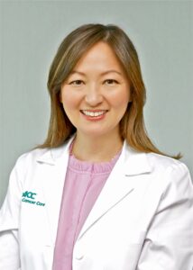 Smiling woman in a white lab coat with NCC Cancer Care logo, pink blouse, portrait photo, professional medical provider.
