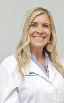 Smiling blonde woman in a white lab coat with a light blue shirt, Atlanta Cancer Care logo visible on the coat, neutral gray background. 